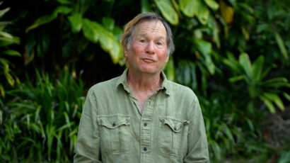 A man in a light green shirt with foliage in the background.