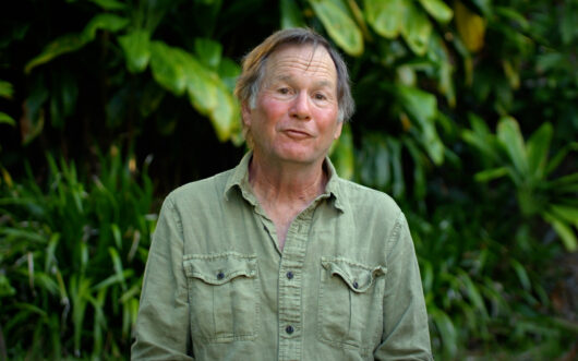 A man in a light green shirt with foliage in the background.