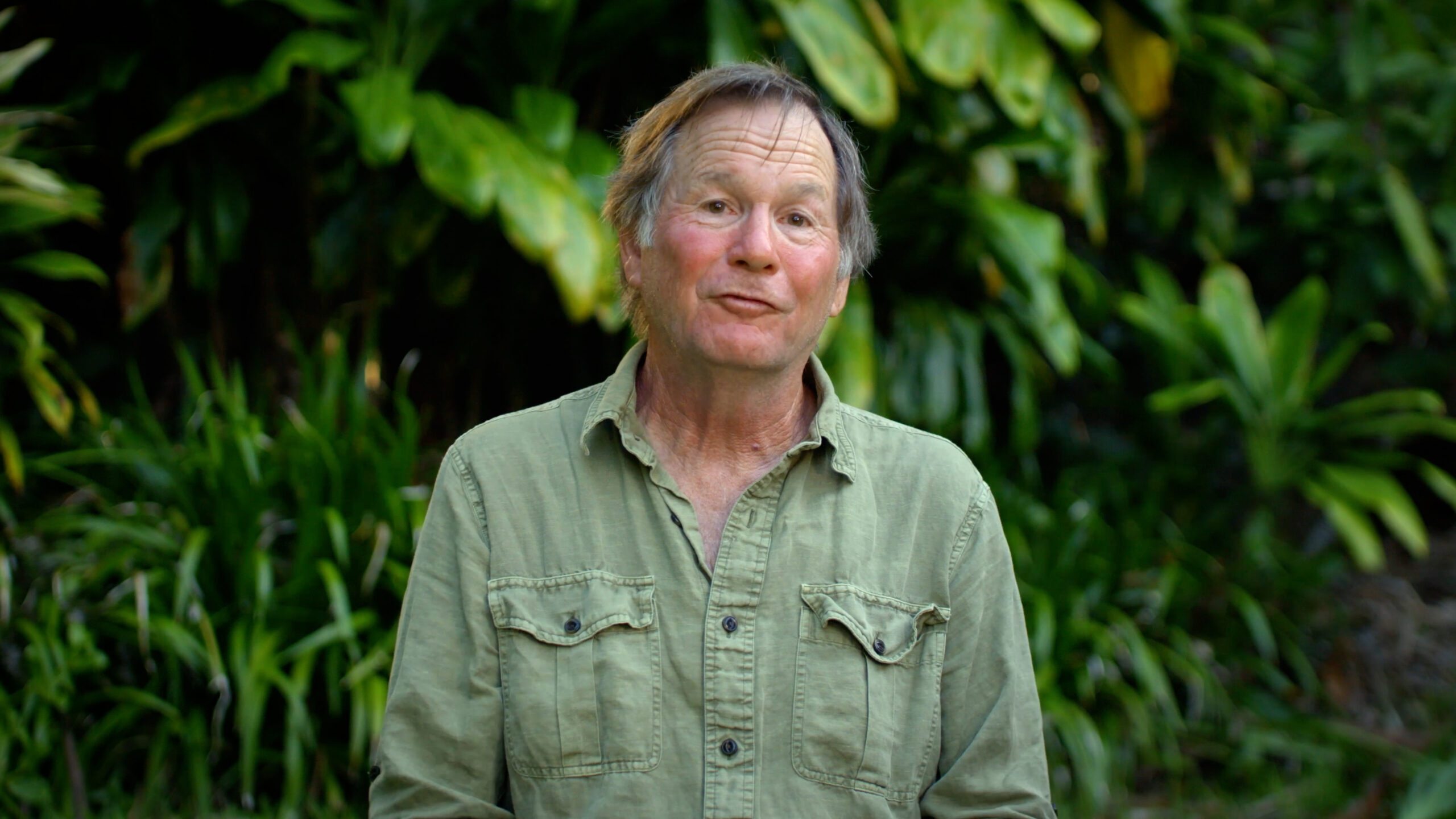 A man in a light green shirt with foliage in the background.