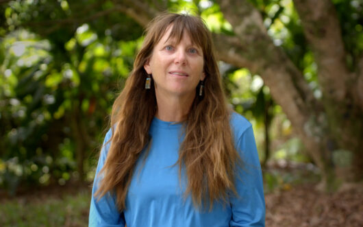 A woman with long brown hair and a blue shirt standing in front of a tree.