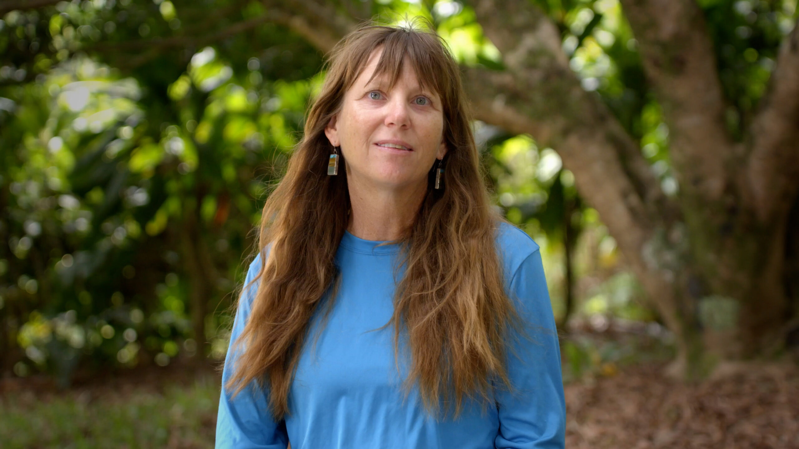 A woman with long brown hair and a blue shirt standing in front of a tree.