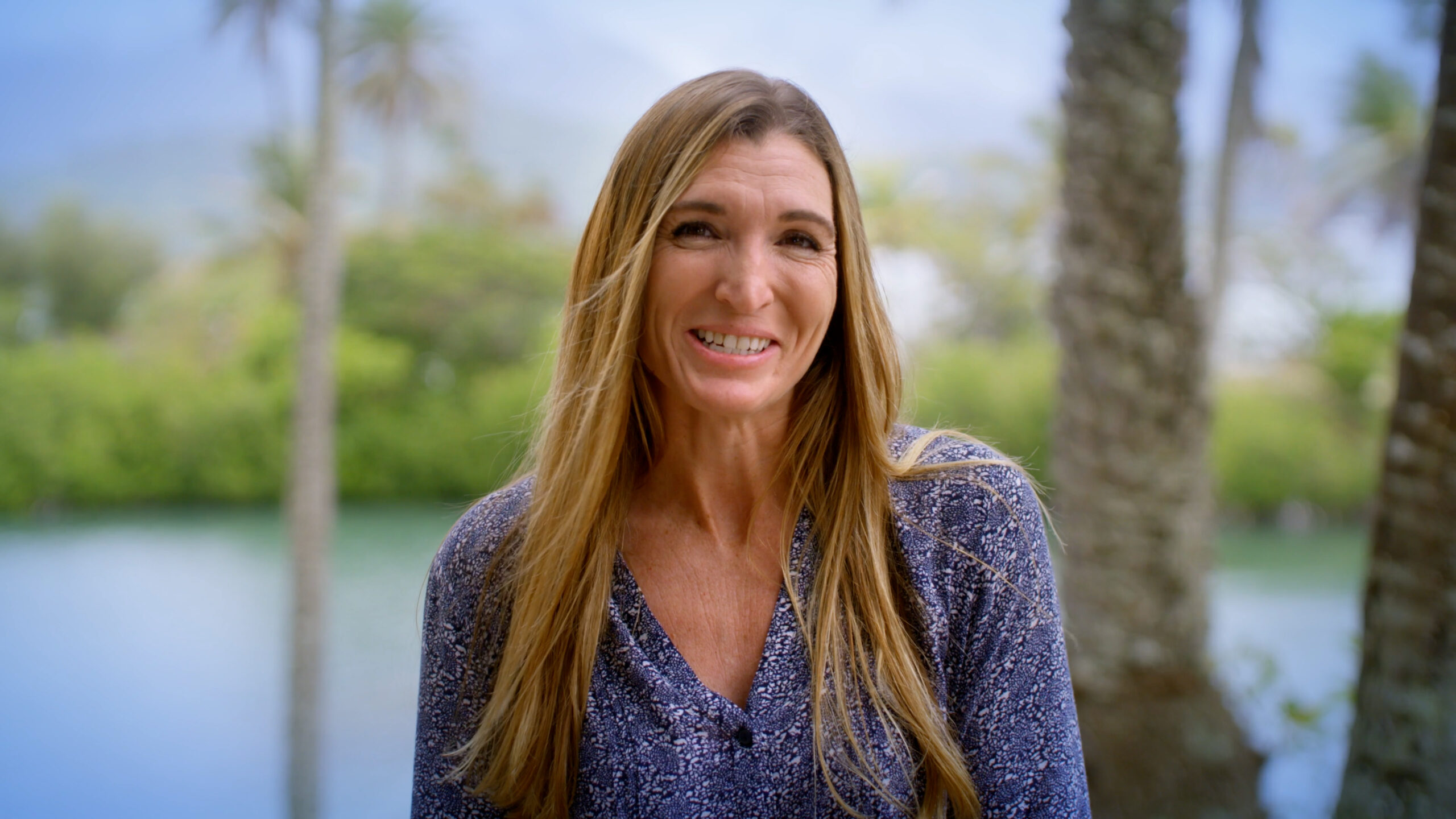 A woman with long blonde hair looking at the camera in front of a tropical background.