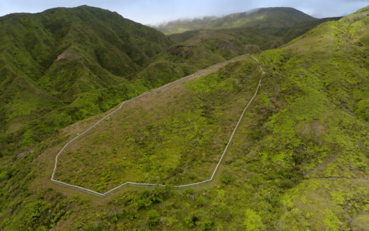 A fence on a green hillside with two more green hills in the backgorund.