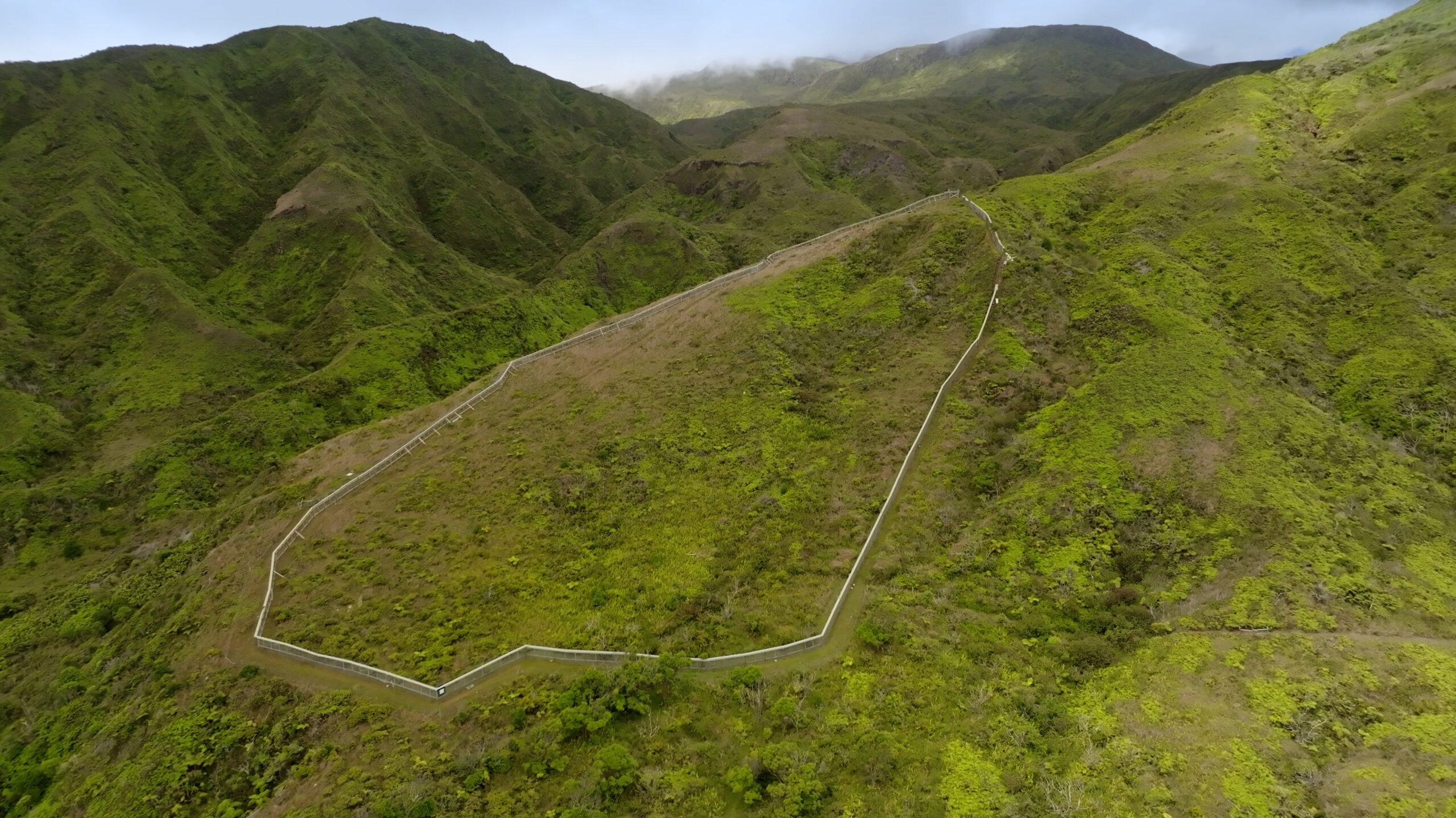 A fence on a green hillside with two more green hills in the backgorund.