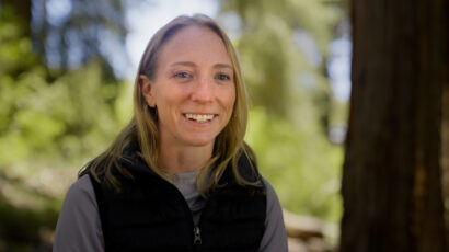 A blonde woman in a forest smiles at the camera.