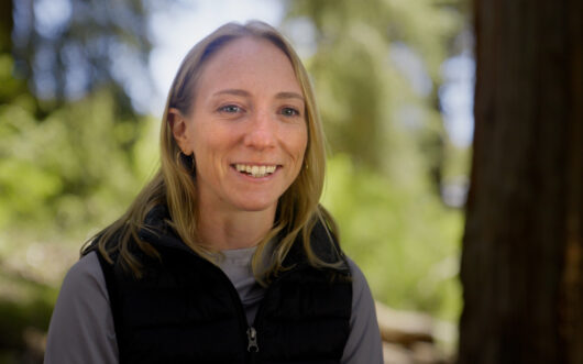 A blonde woman in a forest smiles at the camera.