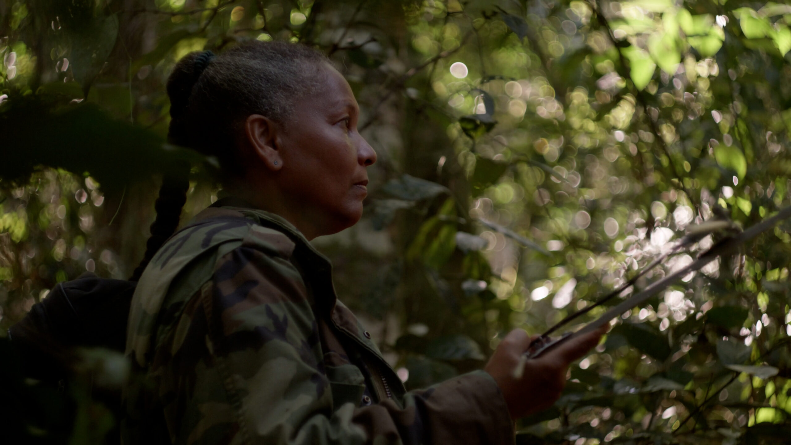 A woman with a green shirt standing in profile, in the shade of a forest.