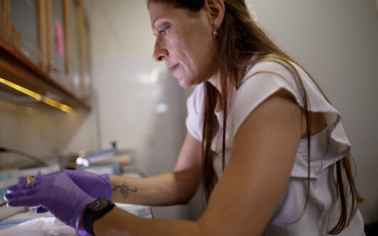 A woman with long brown hair wearing a white t-shirt looks a petri dish.