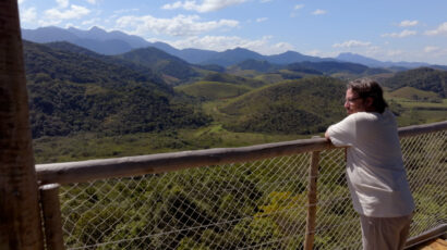 A man in a white t-shirt leans on a fence, looking out over a forested mountain landscape.