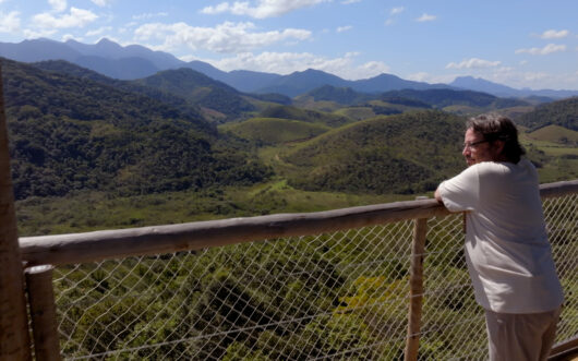 A man in a white t-shirt leans on a fence, looking out over a forested mountain landscape.