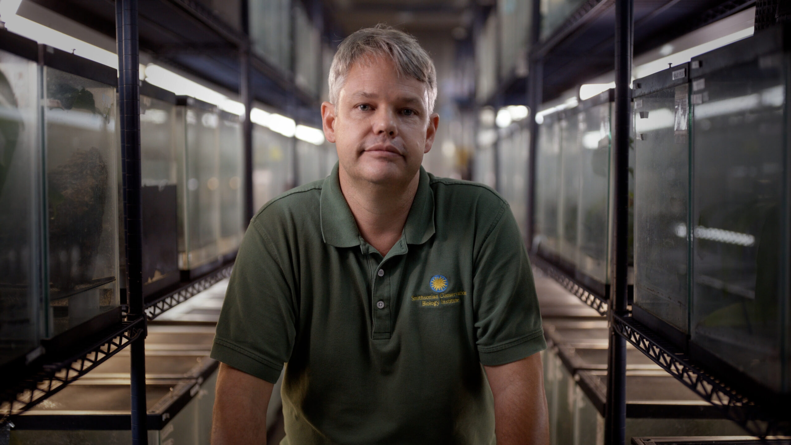 A man in a green t-shirt with gray hair sits in front of a hallway.