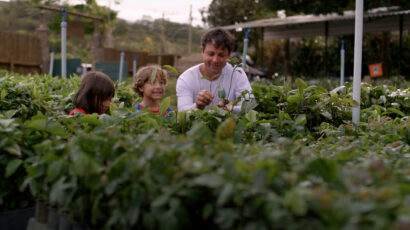 A man in a white shirt and two children stand in a plant nursery surrounded by saplings.