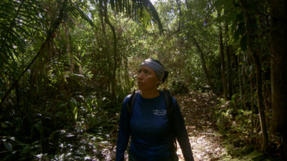 A woman walks in a shadowy rainforest.